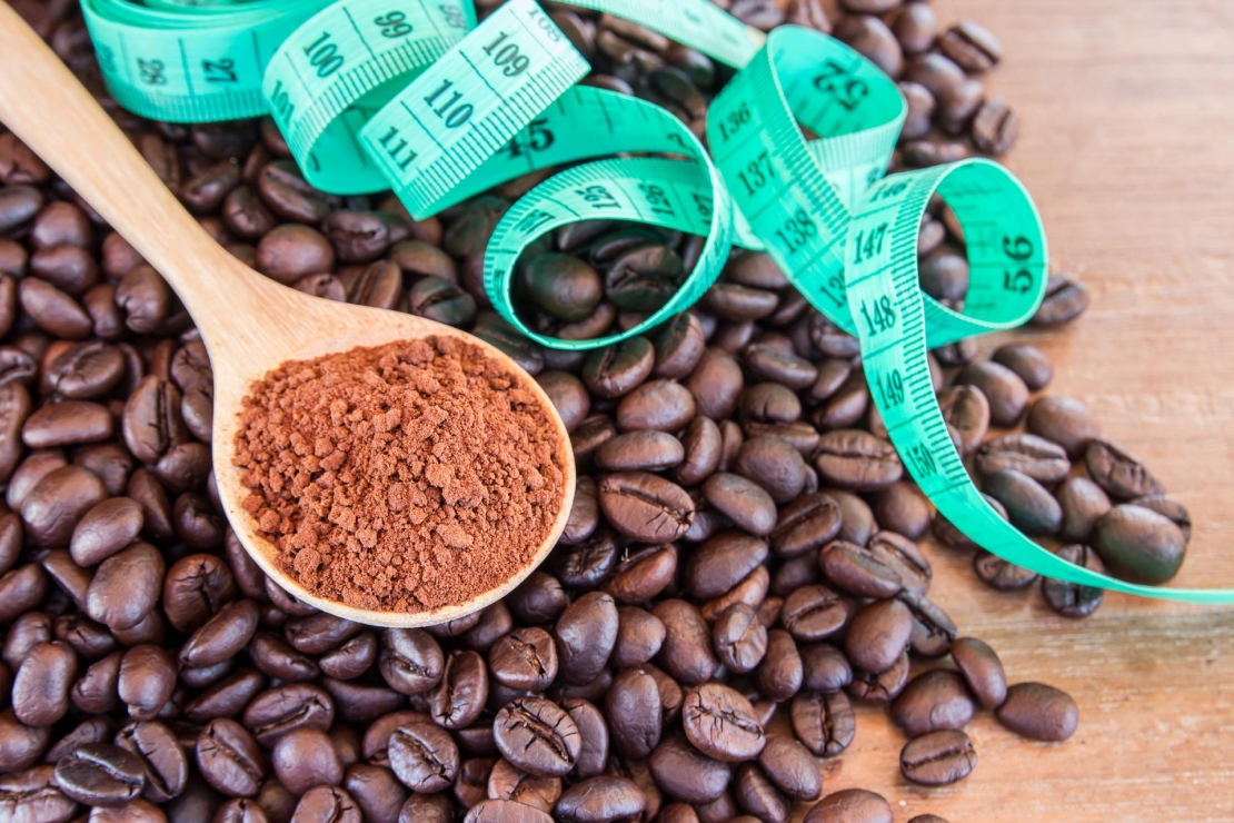 A measuring tape wrapped around a pile of coffee beans with a wooden spoon holding ground coffee, symbolizing the link between coffee, measurement, and fat loss.